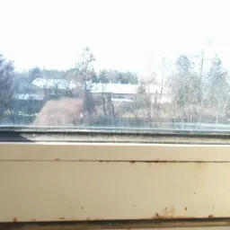 Brudne okno z widokiem na drzewa i budynki w tle, widoczne zacieki i kurz na szybie oraz zabrudzenia na ramie okiennej.