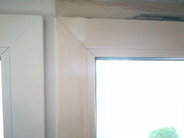 Fragment ramy okiennej z widocznymi śladami po uszczelnianiu, z delikatnym zarysem zieleni za oknem.