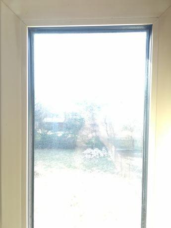 Zabrudzone okno z widokiem na zewnątrz, przez które przebija silne światło słoneczne, ukazujące zarys ogrodu z krzewami i trawnikiem.