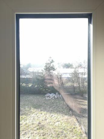 Widok z okna na zewnątrz: trawnik, drzewa i budynki w oddali, widoczne zabrudzenia na szybie.