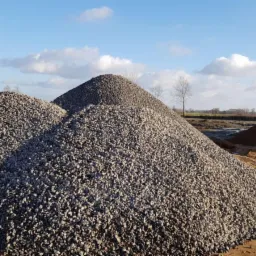 Trzy kopce szarego kruszywa o różnej wielkości na placu budowy, widoczne ślady opon na piasku, błękitne niebo z obłokami w tle.