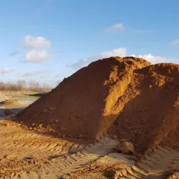 Góra sypkiego, brązowego materiału z widocznymi śladami opon pojazdów na powierzchni, skąpana w popołudniowym słońcu na tle błękitnego nieba z chmurami.