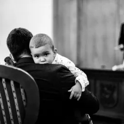 Czarno-białe zdjęcie z ceremonii: ojciec trzyma na rękach małego chłopca, który patrzy w obiektyw zza jego ramienia, w tle widoczny urzędnik.