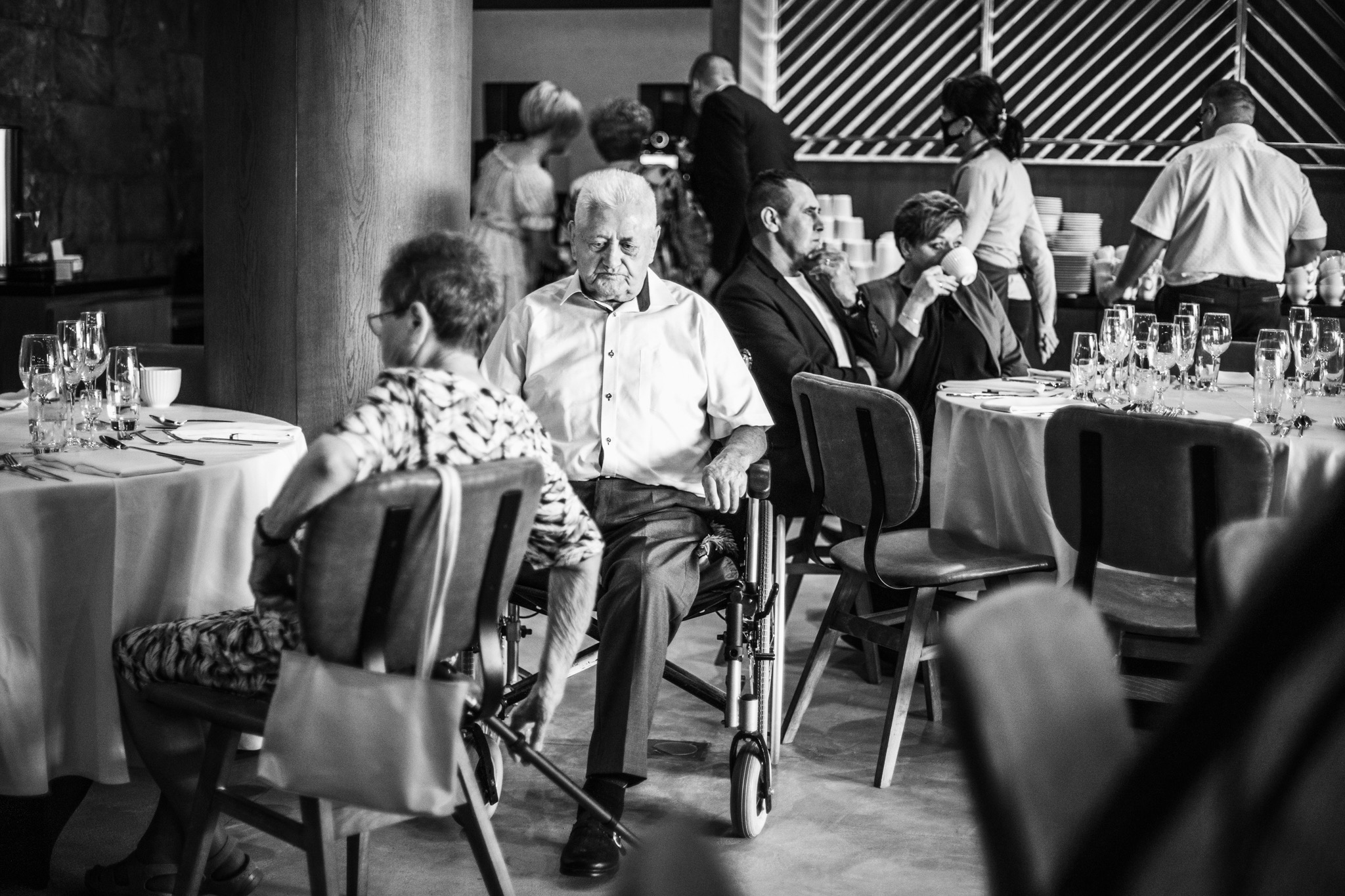 Czarno-białe zdjęcie gości siedzących przy stołach podczas uroczystości, z mężczyzną na wózku inwalidzkim na pierwszym planie.