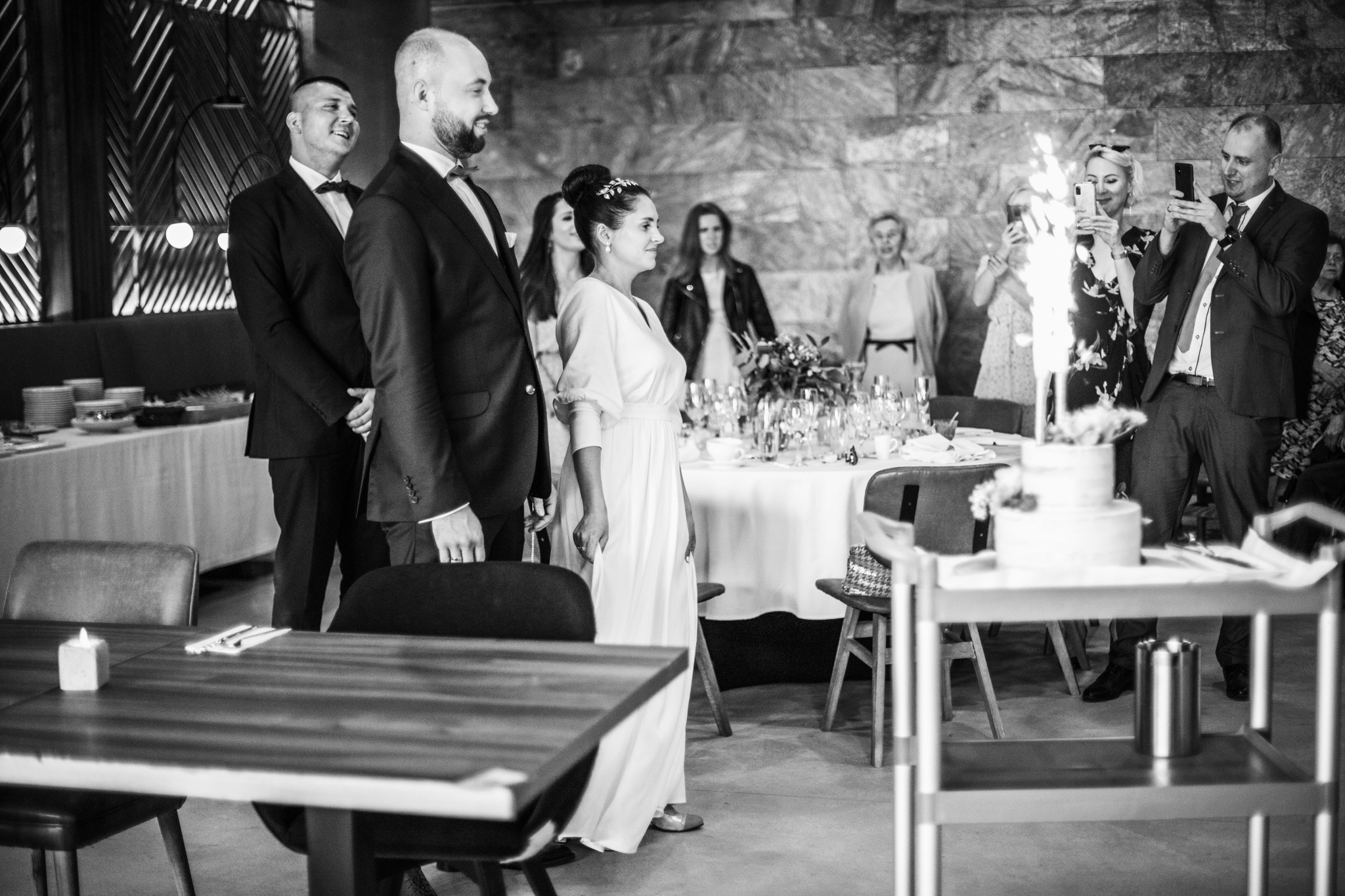 Czarno-białe zdjęcie pary młodej stojącej obok siebie, goście robią zdjęcia płonących zimnych ogni na torcie weselnym, w tle dekoracje i nakryty stół.