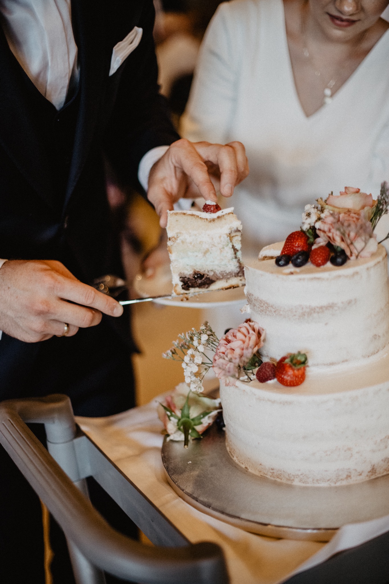 Para kroi trzypiętrowy tort weselny z kremem, ozdobiony kwiatami i owocami, na metalowym stojaku.