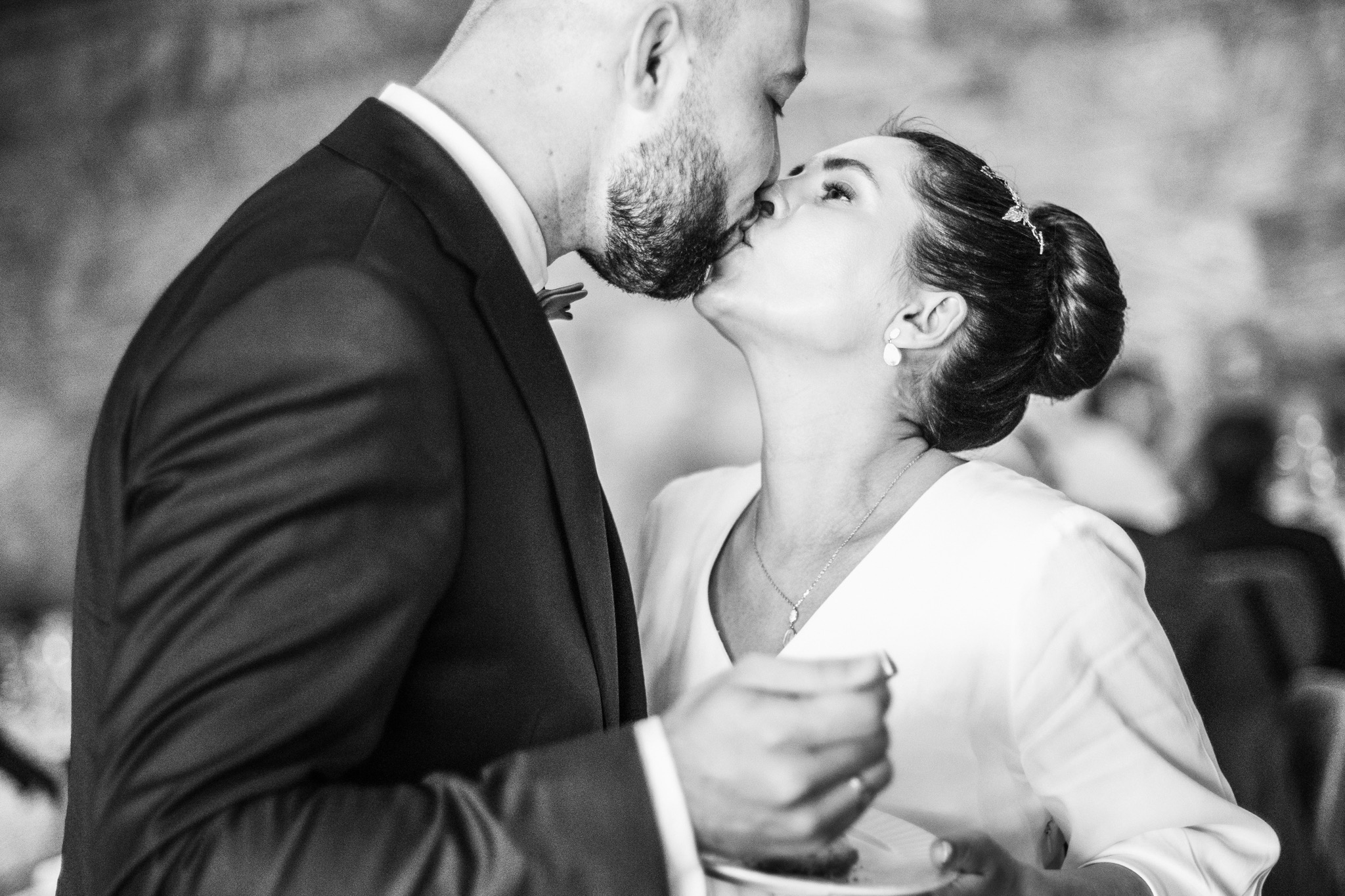 Czarno-białe zdjęcie całującej się pary młodej: on w ciemnym garniturze z muszką, ona w białej sukience z upiętymi włosami i ozdobną spinką.