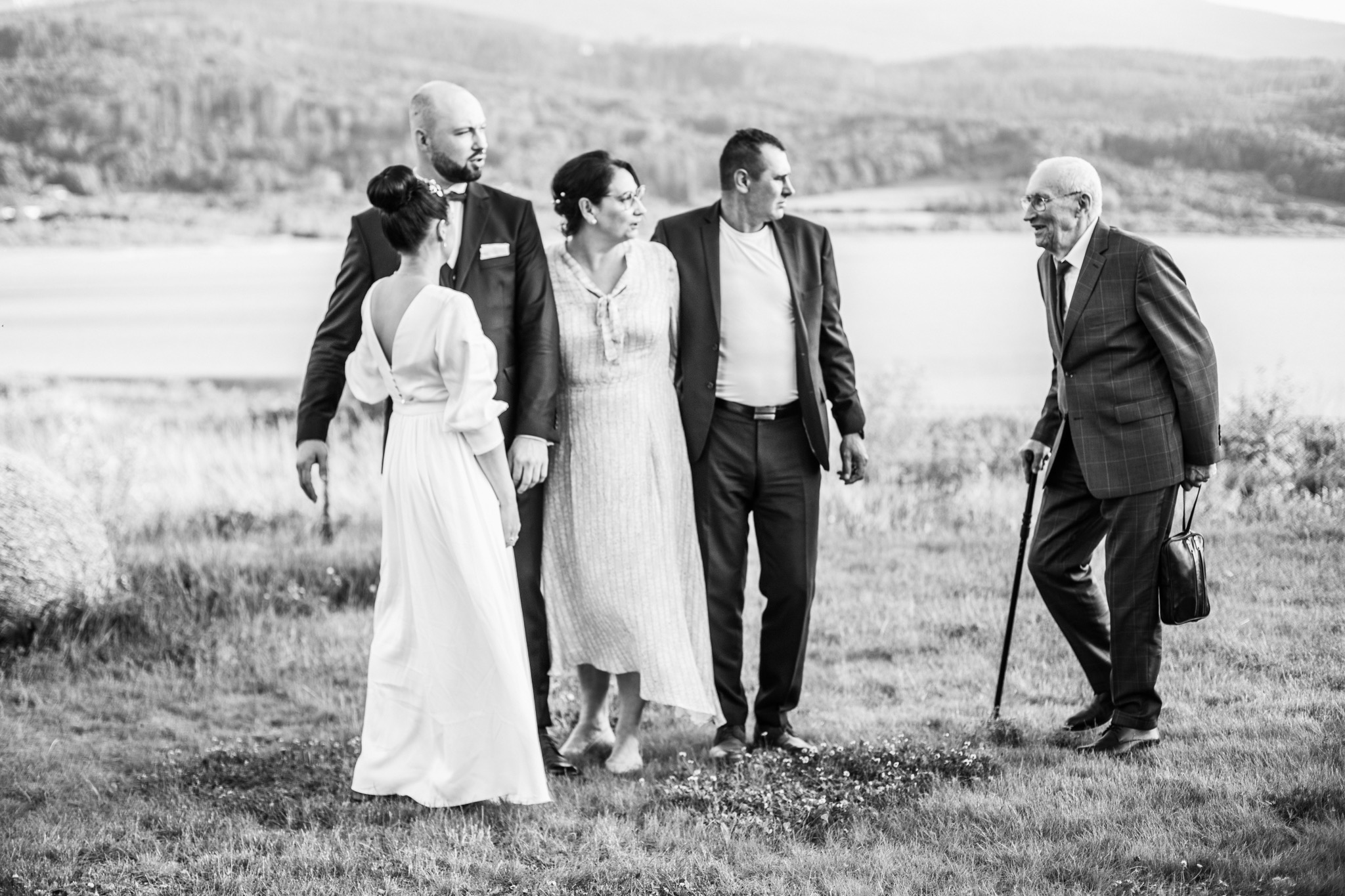 Czarno-białe zdjęcie przedstawia rodzinę na tle jeziora i wzgórz: panna młoda w sukni z odkrytymi plecami, pan młody w smokingu, kobieta w sukience do kolan i mężczyzna w garniturze patrzą w stronę...