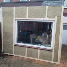 Ocieplona ściana zewnętrzna z zamontowanym oknem, widoczna warstwa izolacyjna z wełny mineralnej, drewniany szkielet konstrukcyjny, ceglana podmurówka i kostka brukowa na ziemi.