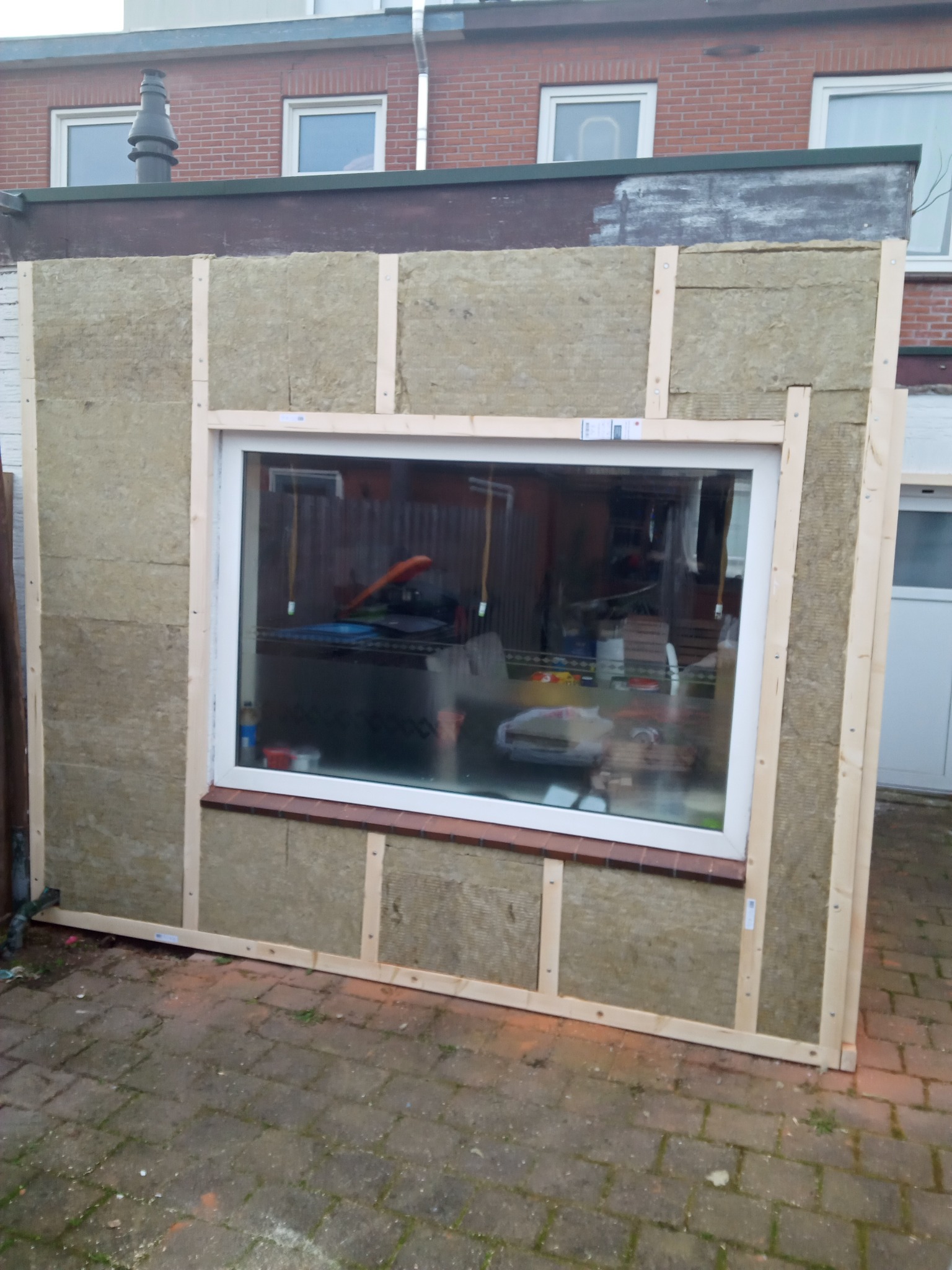 Ocieplona ściana zewnętrzna z zamontowanym oknem, widoczna warstwa izolacyjna z wełny mineralnej, drewniany szkielet konstrukcyjny, ceglana podmurówka i kostka brukowa na ziemi.