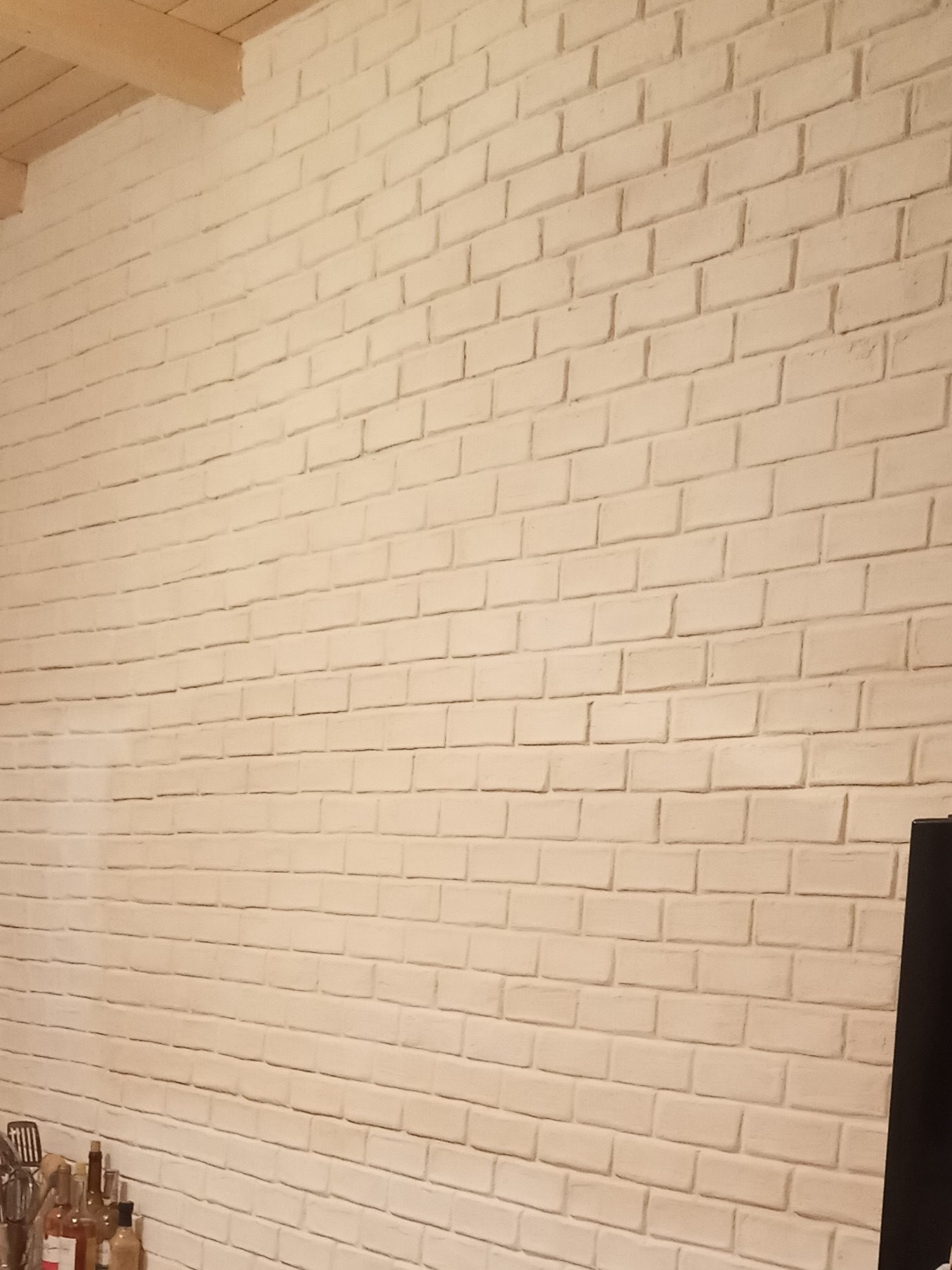 Ściana z białej cegły w pomieszczeniu, z widoczną drewnianą belką sufitową i fragmentem wyposażenia kuchni w dolnym lewym rogu.