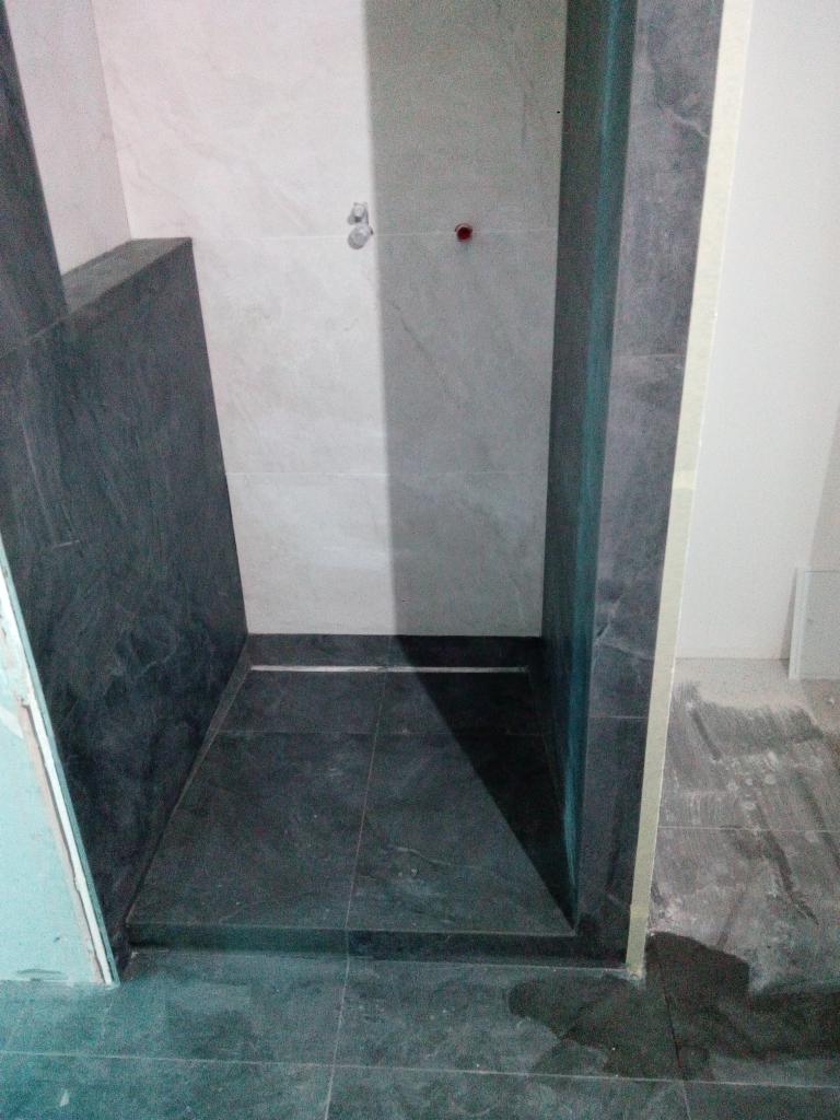 Prysznic w trakcie wykańczania: ciemne płytki podłogowe i ścienne, widoczne punkty instalacji wodnej, surowe krawędzie ścian.