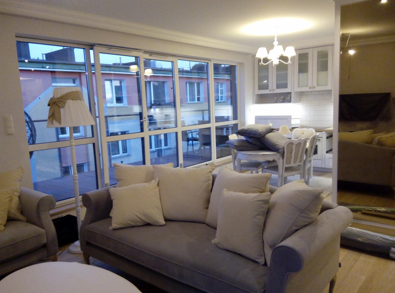 Jasne wnętrze mieszkania z szarym narożnikiem z poduszkami, widok na balkon z dużymi oknami i zabudowaną kuchnię w tle.