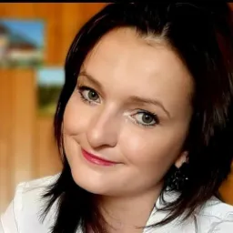 Portret uśmiechniętej kobiety z ciemnymi włosami i jasnymi oczami, ubrana w białą koszulę, z rozmytym tłem.