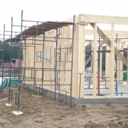 Wznoszenie konstrukcji drewnianej domu na fundamencie, widoczne rusztowania i elementy instalacji, teren budowy w trakcie prac.