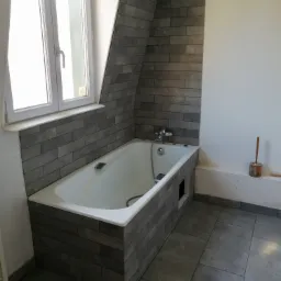 Biała wanna obudowana szarymi płytkami imitującymi kamień, z baterią i słuchawką prysznicową, w łazience wykończonej w szarościach, widok na okno i ukośną ścianę.