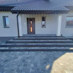 Nowoczesny dom jednorodzinny z ciemną dach&oacute;wką i szarą elewacją, z wejściem wyłożonym kostką brukową w odcieniach szarości, prowadzącym po czterech stopniach do drzwi wejściowych