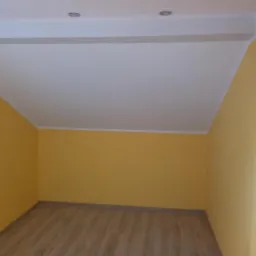 Pomalowane na żółto wnętrze pokoju na poddaszu ze skośnym sufitem i panelami na podłodze, widoczne dwa reflektory w suficie.