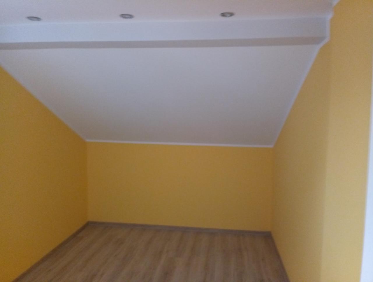 Pomalowane na żółto wnętrze pokoju na poddaszu ze skośnym sufitem i panelami na podłodze, widoczne dwa reflektory w suficie.