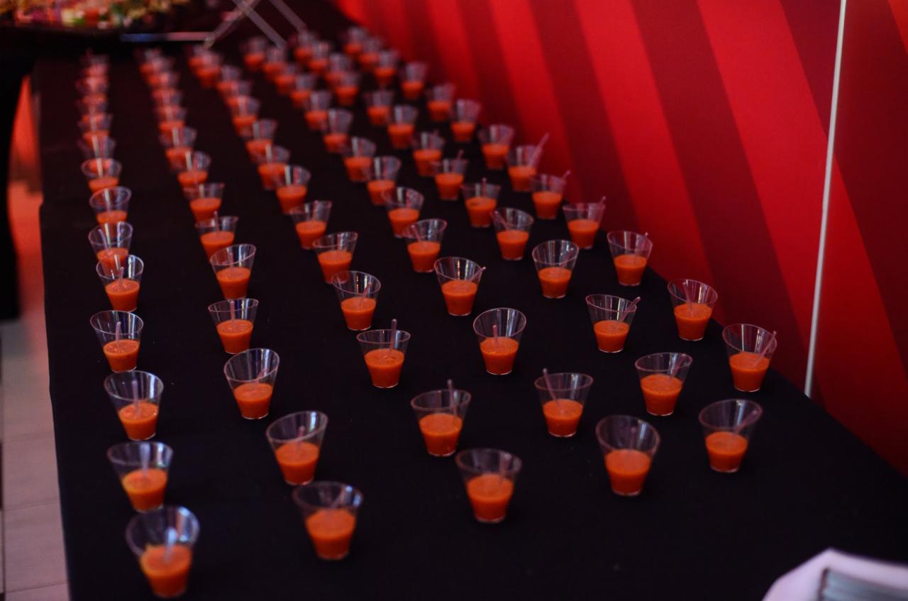 Rzędy małych, plastikowych kieliszków wypełnionych pomarańczowym musem na czarnym stole, tło z czerwonymi pasami.