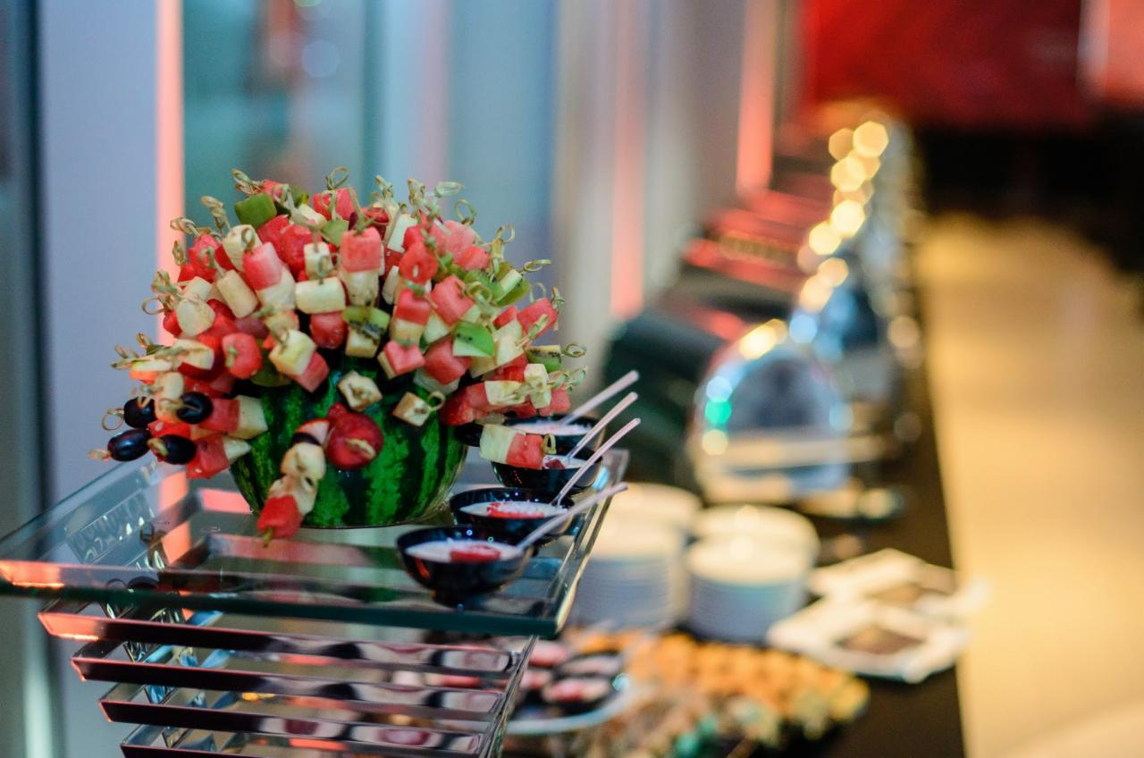 Aranżacja owocowa: arbuz wydrążony i wypełniony szaszłykami z arbuza, kiwi, sera i oliwek, ustawiona na szklanym, wielopoziomowym stojaku obok czarnych miseczek z sosem.