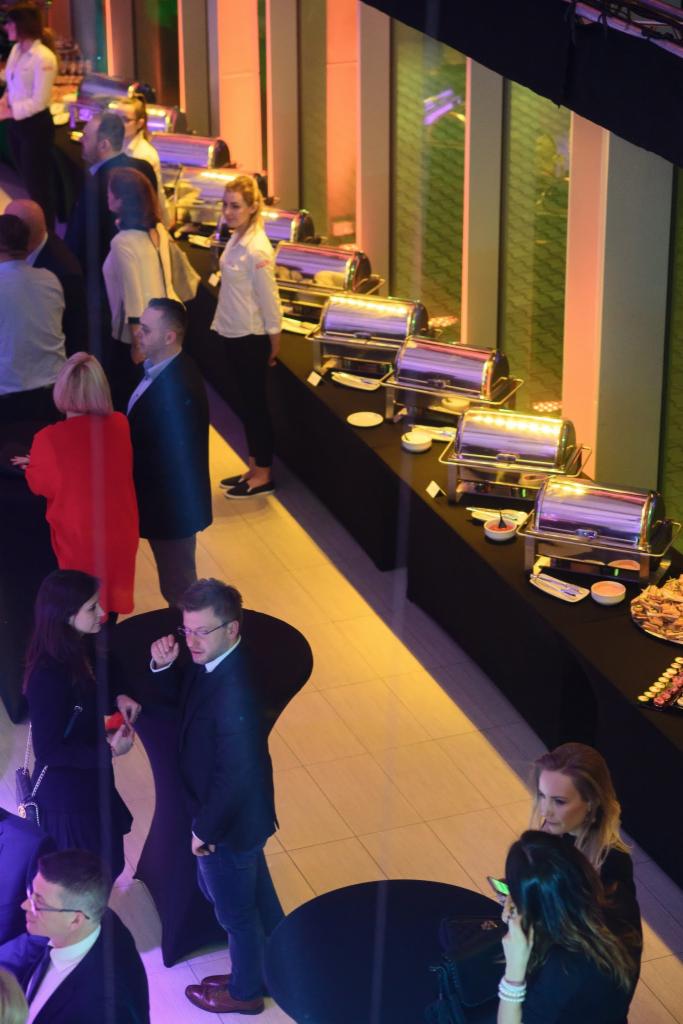 Widok z góry na salę bankietową z bufetem cateringowym, gdzie goście w strojach biznesowych rozmawiają przy stolikach; bufet z podgrzewaczami, naczyniami i przekąskami.