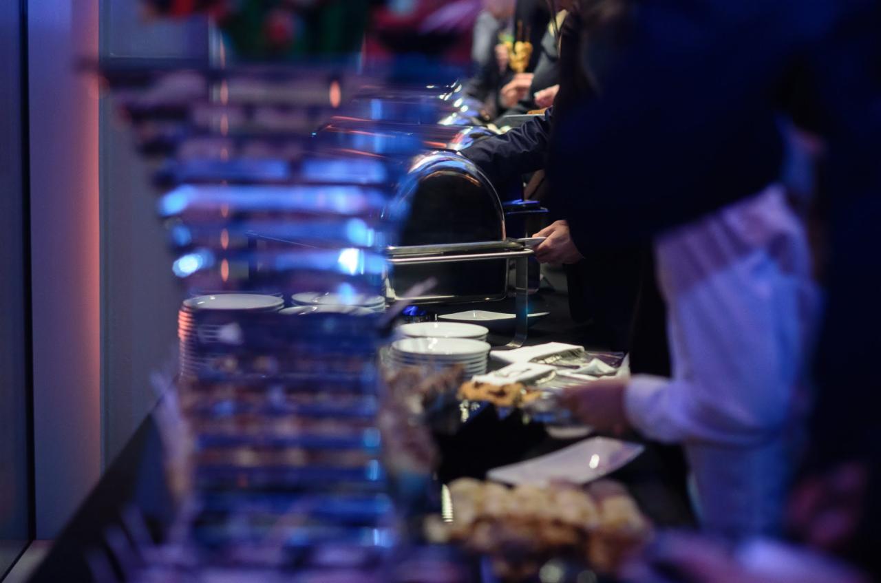 Bufet z podgrzewaczami potraw i stosem talerzy, obsługa w tle, niebieskie oświetlenie