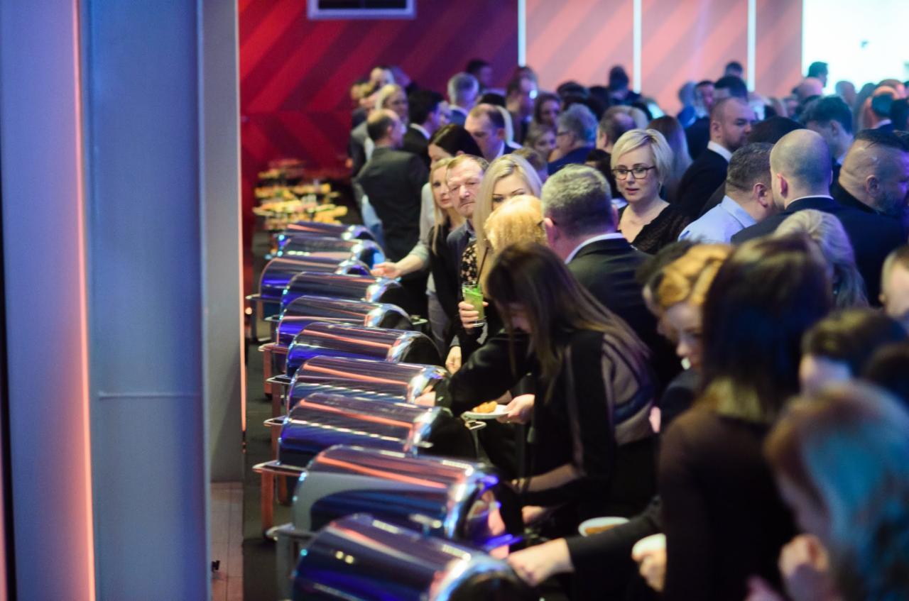 Uczestnicy konferencji w eleganckich strojach, ustawieni w kolejce do bufetu z metalowymi podgrzewaczami, w tle dekoracja w czerwono-białe pasy.
