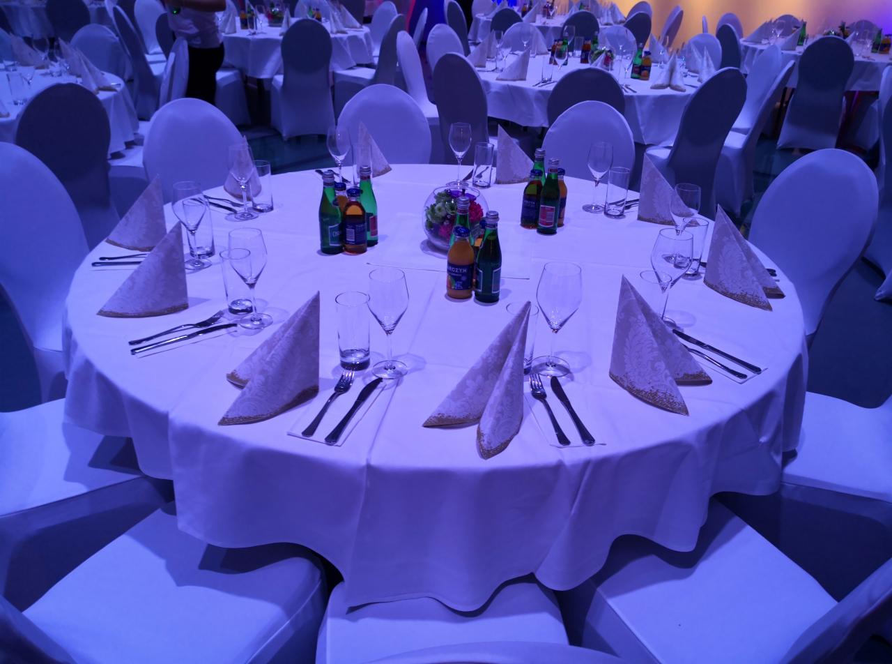 Uroczysty stół z białym obrusem, nakryciami, szklankami i napojami, przygotowany na imprezę firmową lub bankiet, w tle widoczne inne stoły i krzesła.