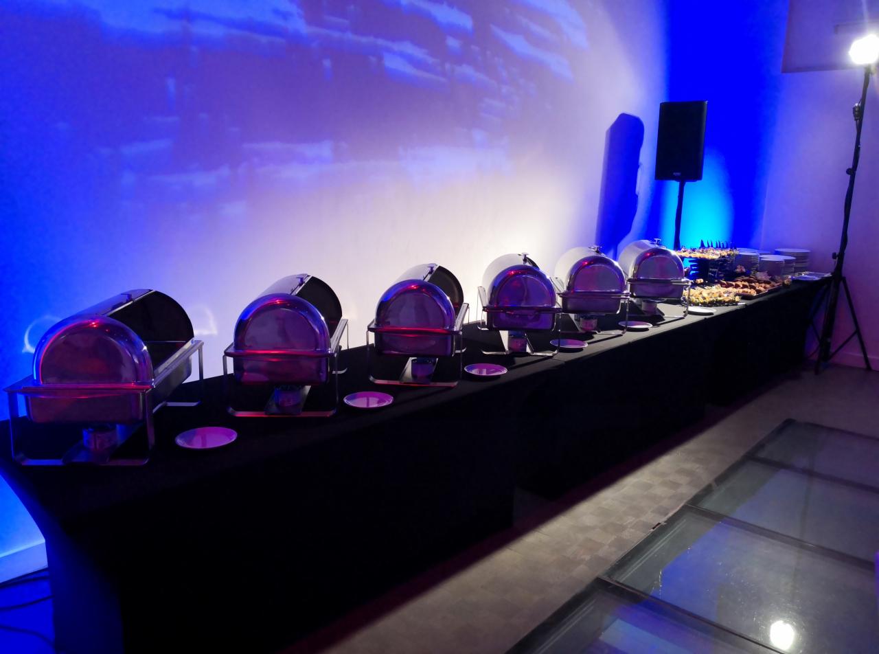 Stół cateringowy z podgrzewaczami, dekorowany czarnym obrusem, oświetlony niebieskim światłem, z widocznymi przekąskami i szklaną podłogą.