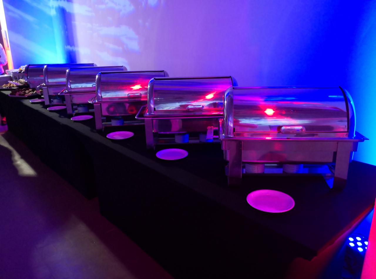 Rząd srebrnych bemarów z potrawami, ustawionych na czarnym stole, oświetlonych niebiesko-fioletowym światłem, z talerzami w kolorze magenta.