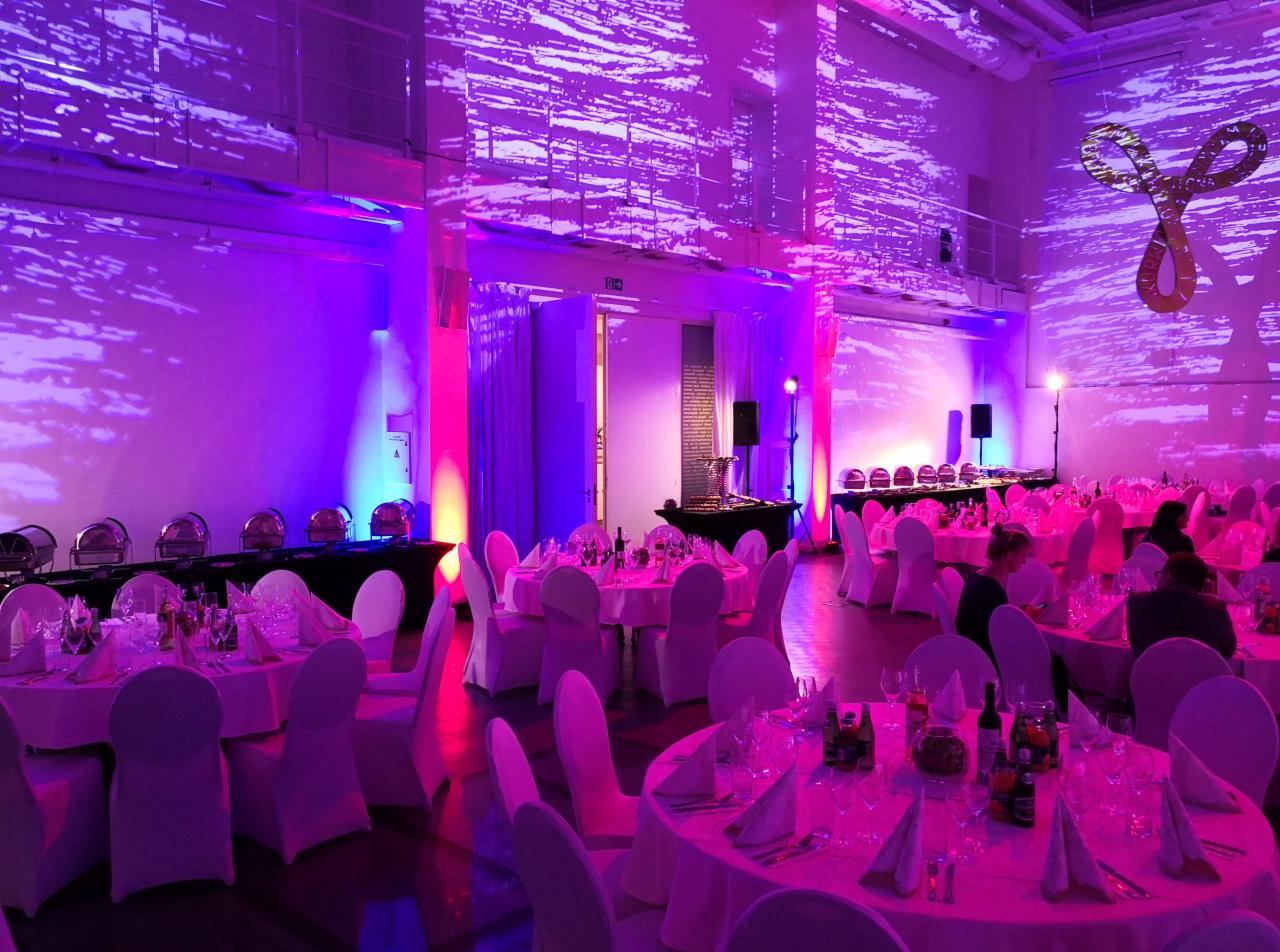 Uroczysta sala bankietowa z okrągłymi stołami nakrytymi białymi obrusami i krzesłami w pokrowcach, oświetlona fioletowym i różowym światłem, z projektorem wyświetlającym wzory na ścianach i bufetem...