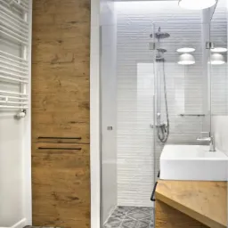 Nowoczesna łazienka z drewnopodobną szafką, kabiną prysznicową ze szklaną ścianką i wzorzystą podłogą w odcieniach szarości oraz białymi lampami wiszącymi.