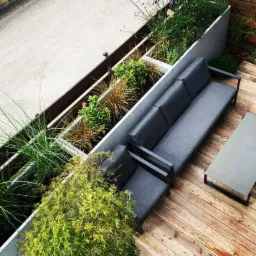 Nowoczesny taras w Rzeszowie z drewnianą podłogą, szarą sofą narożną i podwyższonymi rabatami z różnorodną roślinnością, widok z góry.