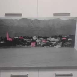 Szklany panel ścienny w kuchni z nadrukiem czarno-białego pejzażu z różowymi akcentami budynków, zamontowany między blatem a szafkami wiszącymi.