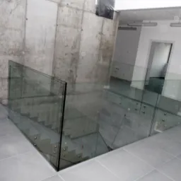 Szklana balustrada zabezpieczająca schody w surowym, betonowym wnętrzu, mocowana punktowo, widoczne odbicia światła w szkle.