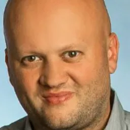 Portret łysiejącego mężczyzny w szarej koszuli na tle błękitnego tła. Widoczna twarz i ramiona, uśmiech, krótko ostrzyżona broda. Neutralny wyraz twarzy.