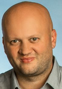 Portret łysiejącego mężczyzny w szarej koszuli na tle błękitnego tła. Widoczna twarz i ramiona, uśmiech, krótko ostrzyżona broda. Neutralny wyraz twarzy.