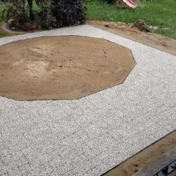 Układanie jasnoszarej kostki brukowej wokół przygotowanego okrągłego placu z piaskiem, widoczne elementy mocujące kostkę, trawnik w tle.