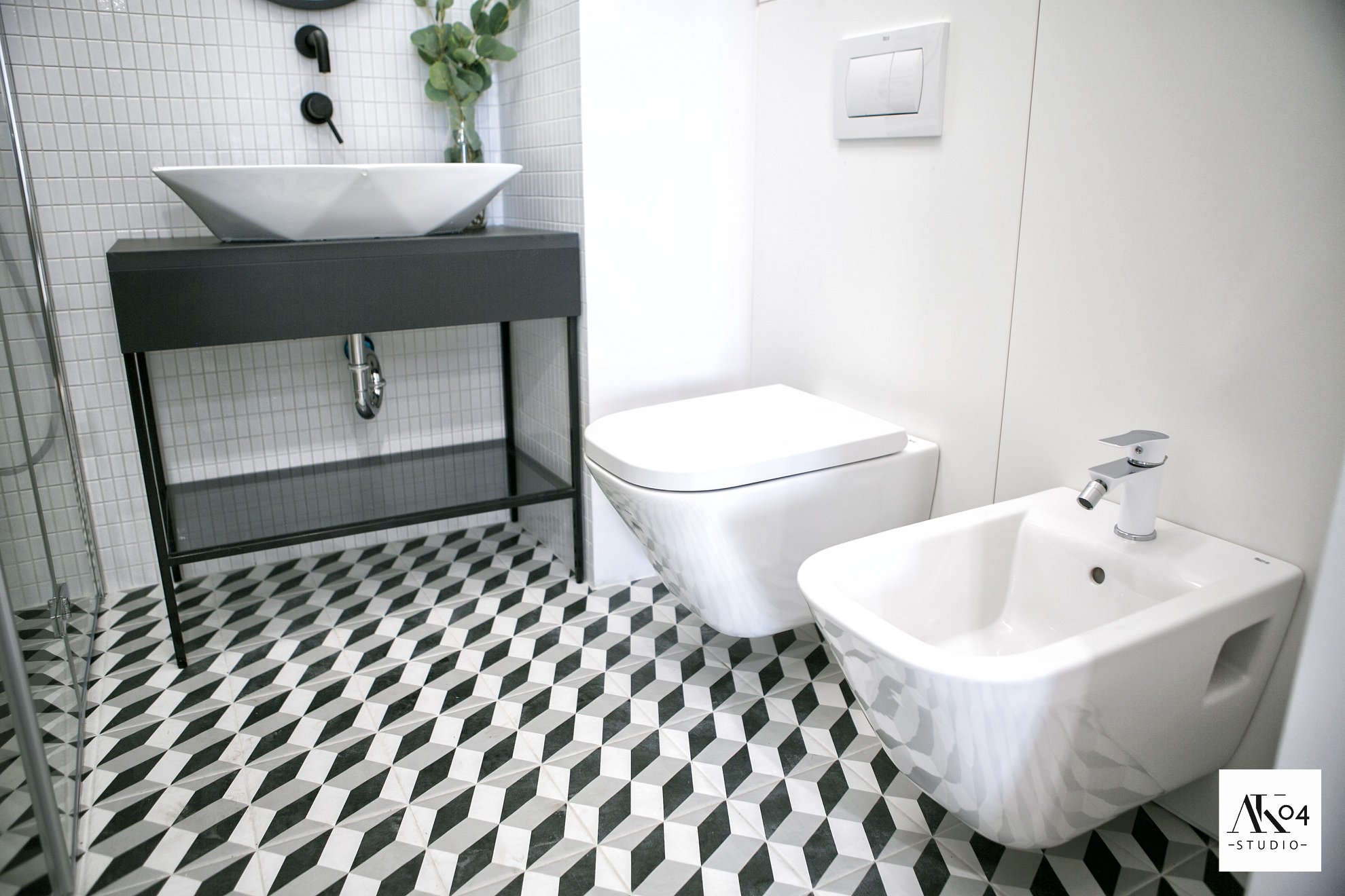 Nowoczesna łazienka z geometryczną podłogą, białą ceramiką sanitarną (umywalka, toaleta, bidet) i czarnymi akcentami, takimi jak bateria umywalkowa i rama umywalki.