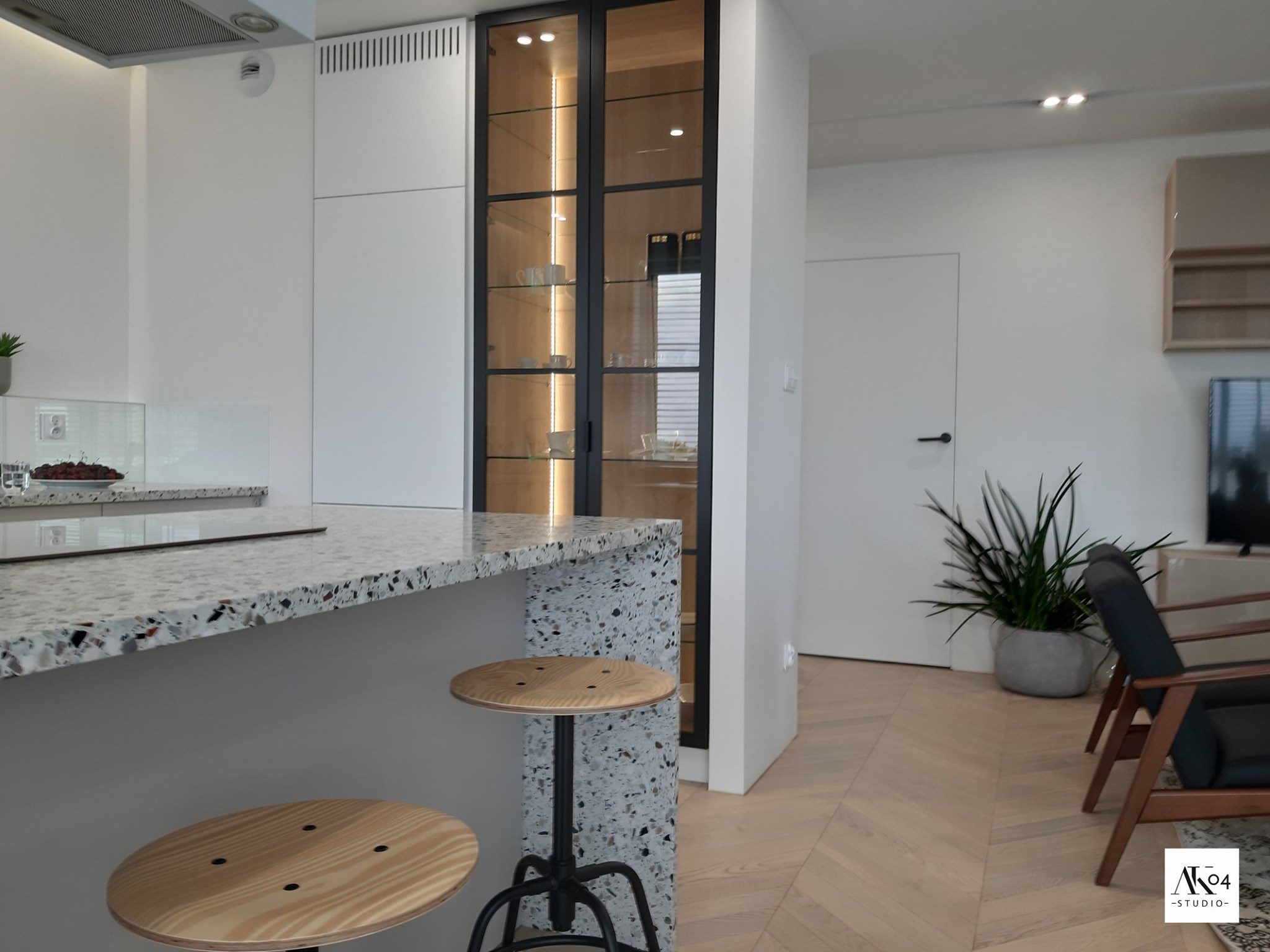 Nowoczesna kuchnia z wyspą wykończoną lastryko, drewniane hokery z czarną podstawą, przeszklona witryna z podświetleniem i porcelaną w tle, parkiet w jodełkę.