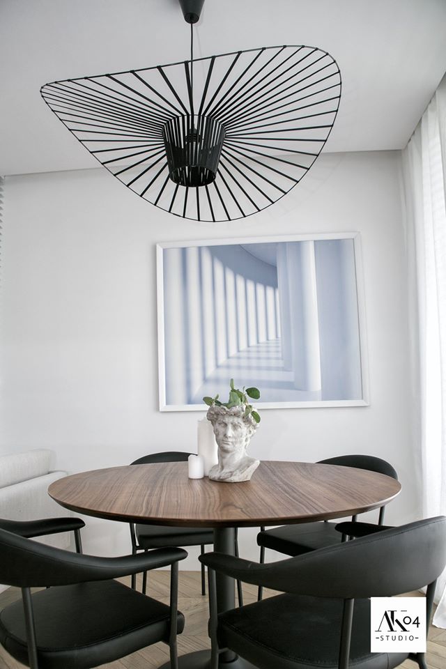 Jadalnia z okrągłym drewnianym stołem, czarnymi krzesłami, nowoczesną lampą wiszącą i dekoracyjną rzeźbą głowy z roślinami.