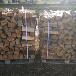 Dwa ułożone stosy drewna opałowego na paletach, zabezpieczone białymi taśmami, widoczne w plenerze, w tle trawa i drzewa.