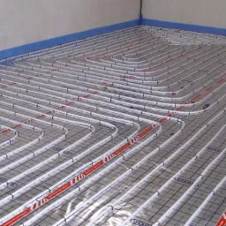 Białe rury ogrzewania podłogowego ułożone równolegle na siatce, zabezpieczone niebieską taśmą przy ścianie, widok z góry.