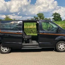 Czarny van z otwartymi drzwiami bocznymi, widoczne siedzenia pasażerskie, pomarańczowy pasek dekoracyjny, tło z zielenią i błękitnym niebem.