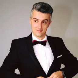 Mężczyzna w czarnym smokingu z r&oacute;żowo-czarną muchą, o siwych włosach, patrzy w lewo, zapinając guzik marynarki.