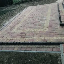 Wykończony taras z kostki brukowej w odcieniach czerwieni i beżu, z dekoracyjnym wzorem obramowania i schodami, otoczony ziemią.