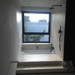 Nowoczesna łazienka z wanną w zabudowie z marmurowym wykończeniem, czarna armatura, okno z widokiem na zieleń, surowe ściany o fakturze betonu.