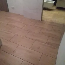 Ułożone płytki podłogowe imitujące drewno w łazience, widoczny fragment toalety i szafki, ściany częściowo przygotowane do malowania.