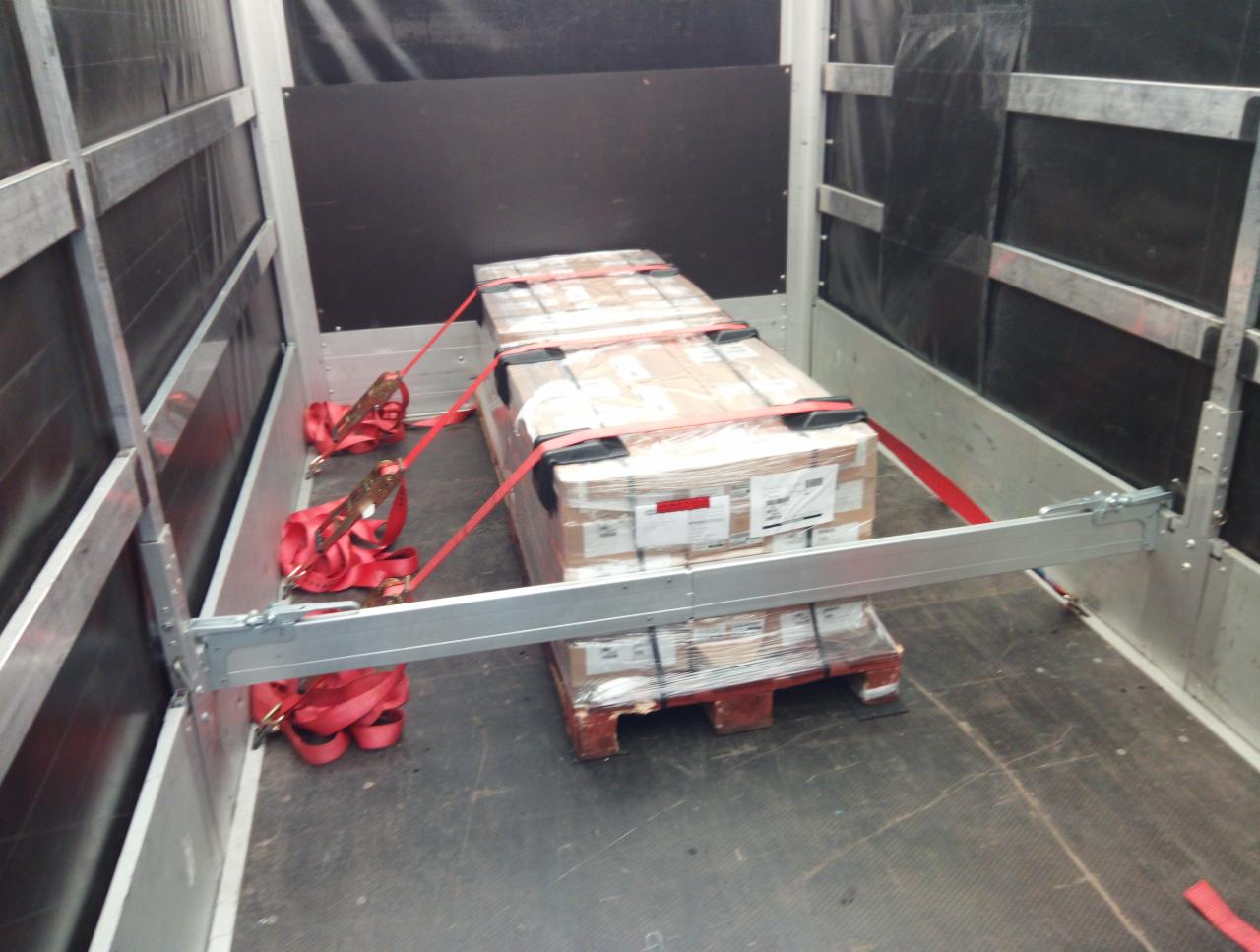 Paleta z kartonami zabezpieczona czerwonymi pasami wewnątrz przestrzeni ładunkowej pojazdu z metalowymi ścianami i podłogą z widocznymi śladami użytkowania.